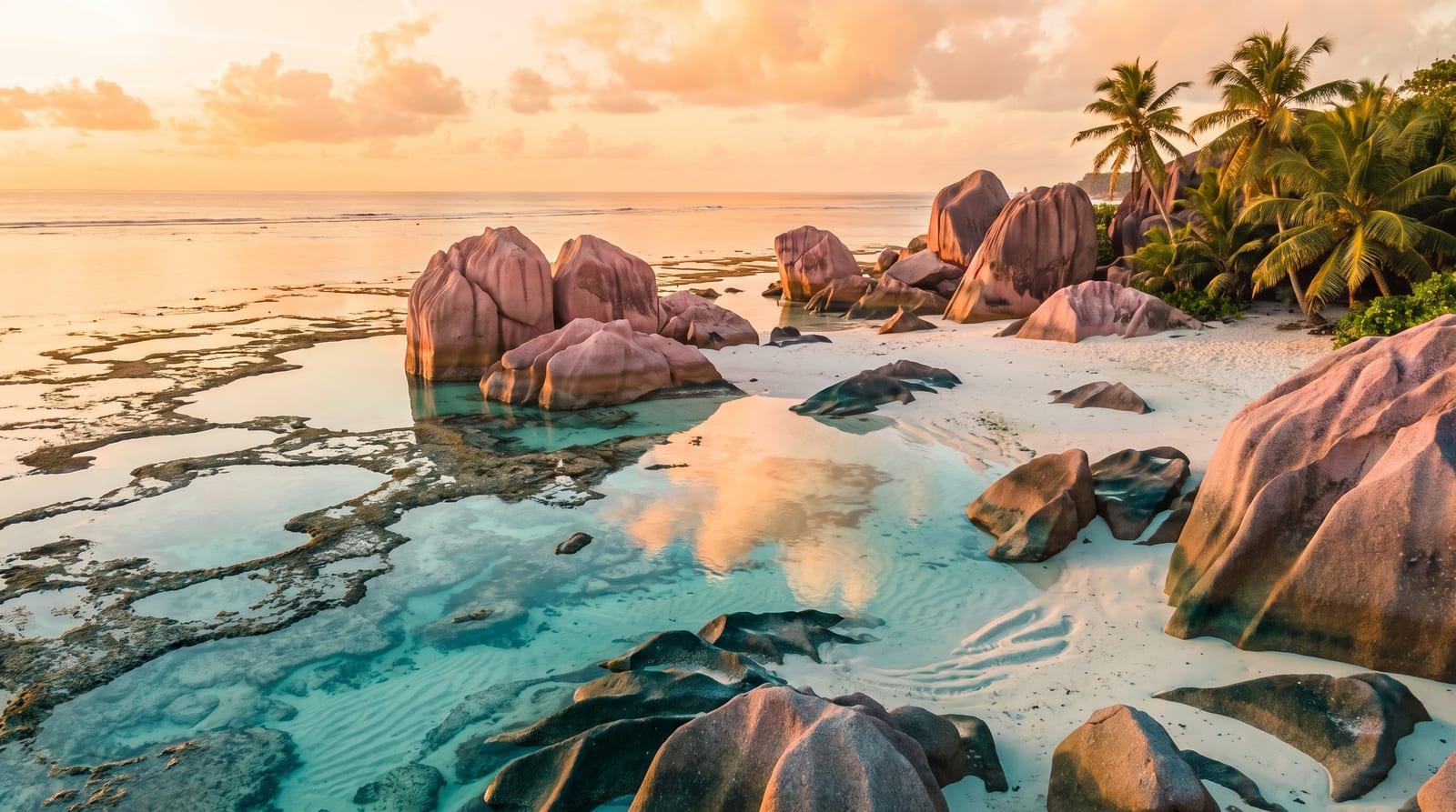 Růžové žulové balvany na pláži Anse Source d'Argent na La Digue za odlivu, mezi nimi mělké tyrkysové tůně odrážející oblohu a kokosové palmy v pozadí.