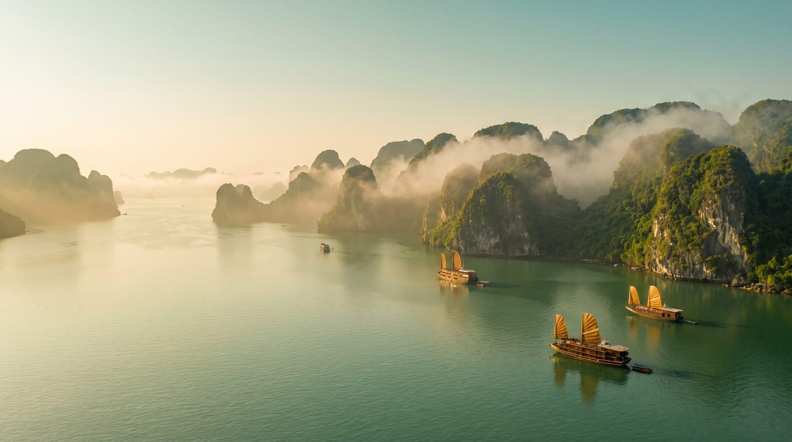 Ranní mlha nad zátokou Ha Long s vápencovými skalami a dřevěnými loděmi na klidné zelené vodě.